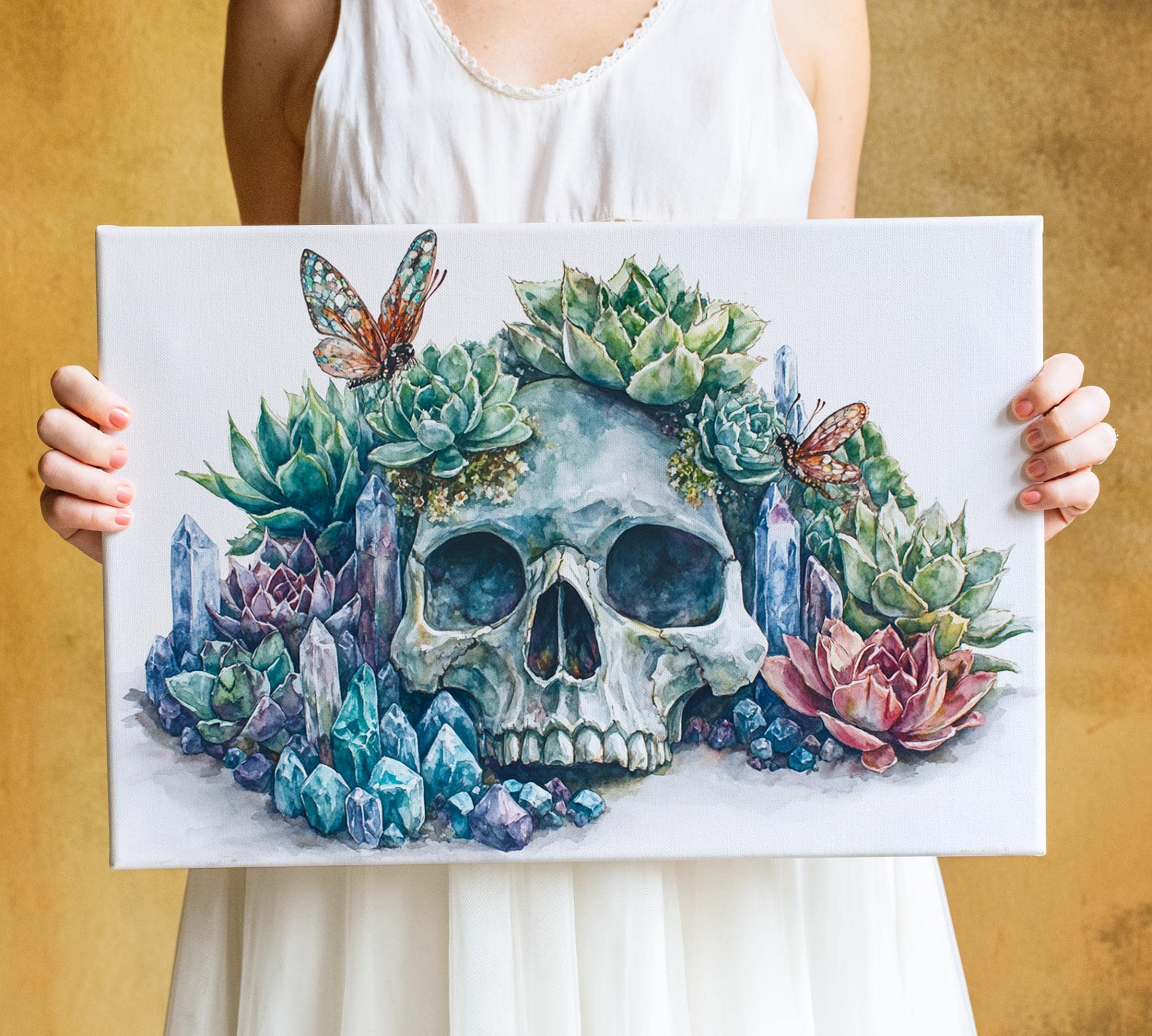 A woman holds a painting of a skull surrounded by a vibrant array of succulents, flowers, and crystals.