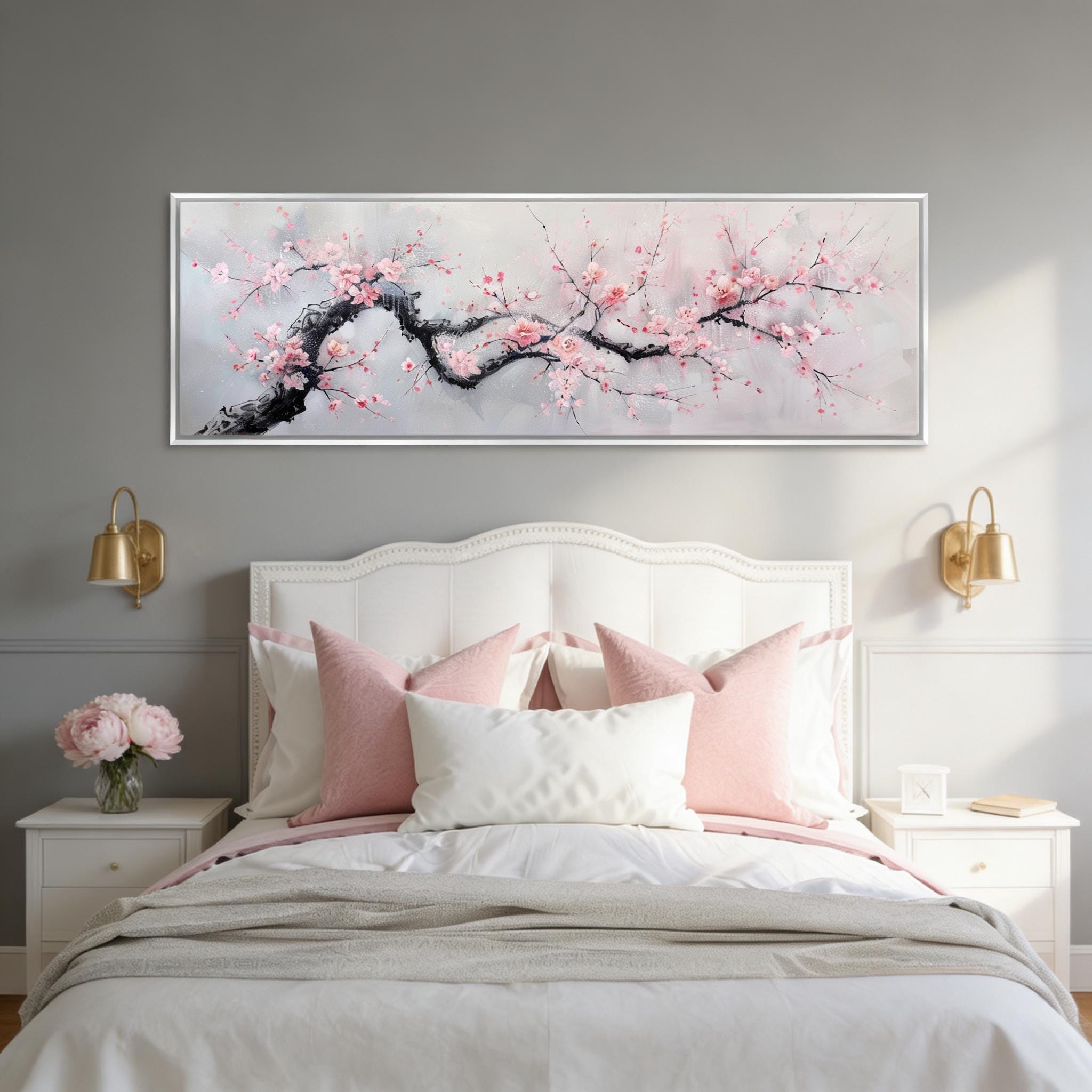 a serene bedroom scene with a large bed adorned with white pillows and a gray blanket, flanked by two white nightstands with gold-colored lamps. Above the bed, a large framed painting of a cherry blossom tree with pink flowers is displayed on the wall.
