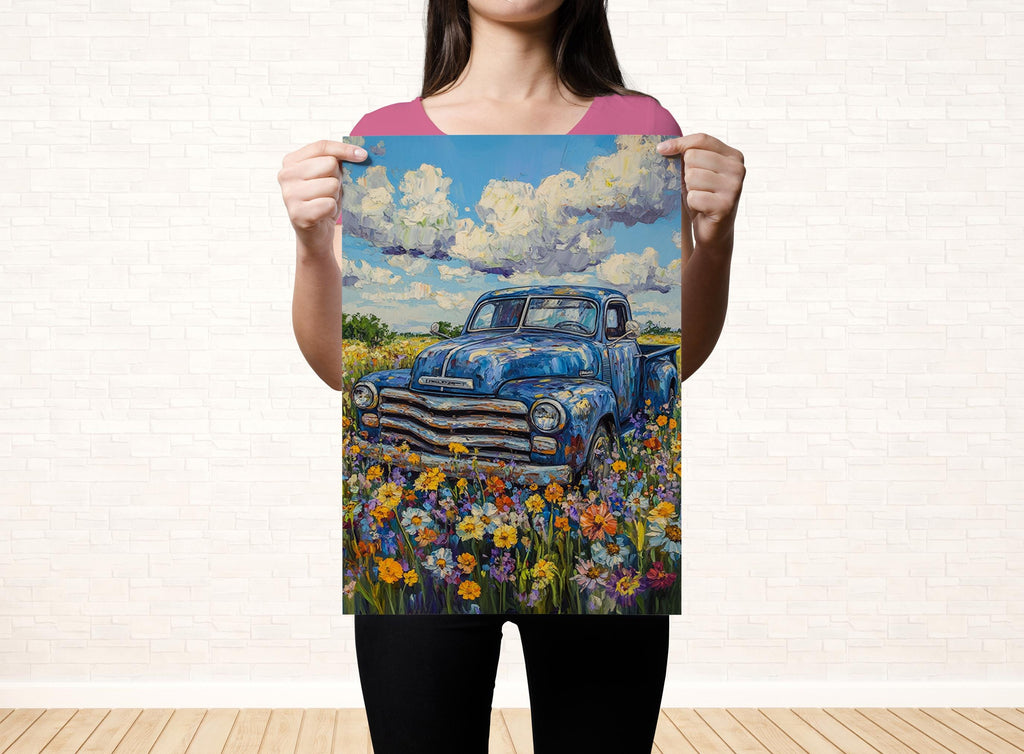 A woman holds up a framed painting of a blue truck parked in a field of colorful flowers.