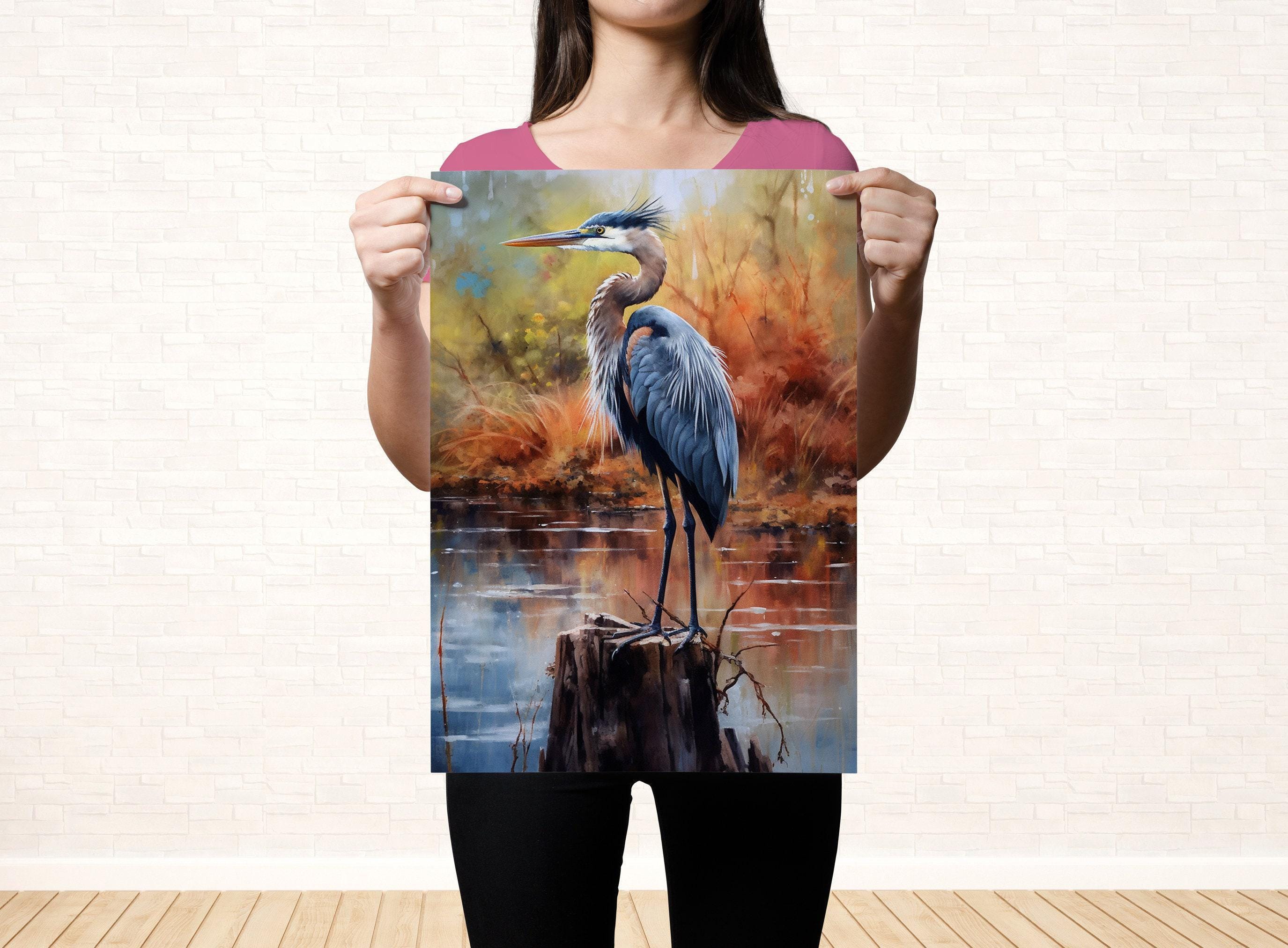 A woman holds up a framed painting of a blue heron standing on a tree stump in a natural setting.