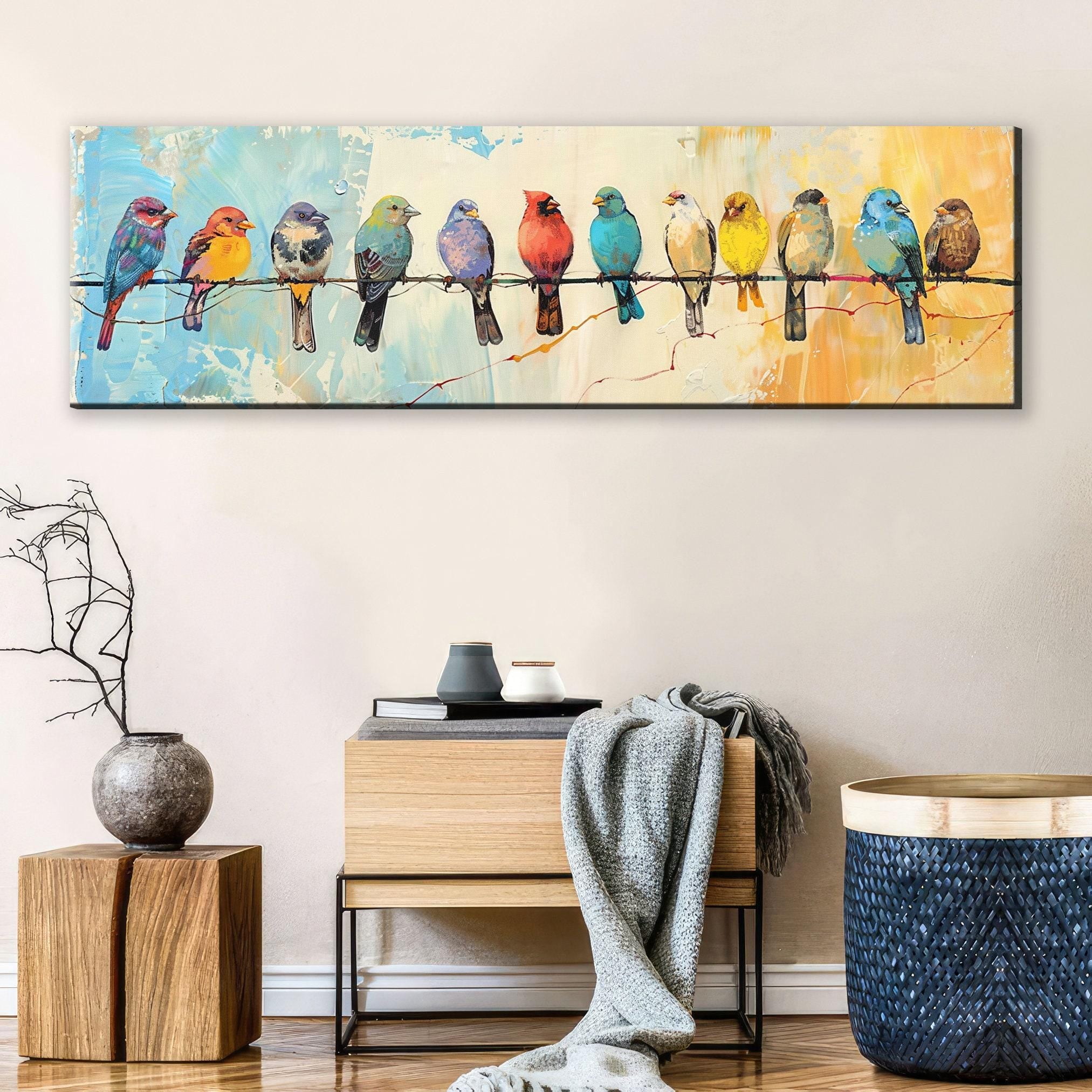 a large painting of colorful birds perched on a wire, displayed on a wall above a wooden table with a vase and a stack of books.