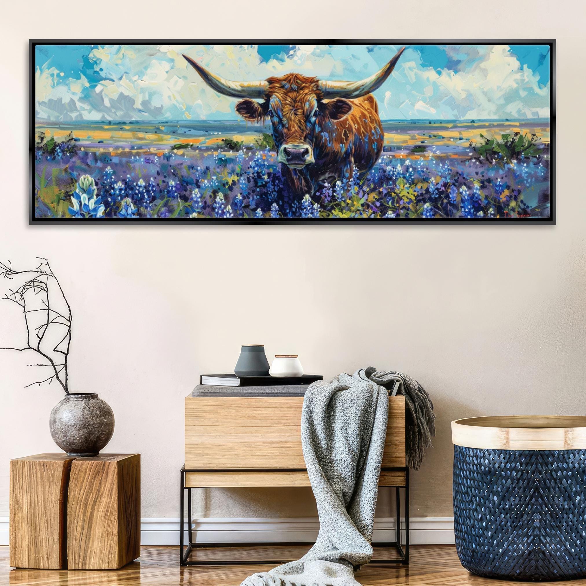 a large painting of a bull with horns, set against a backdrop of a field with purple flowers. The painting is framed and hangs on a wall above a wooden table with a vase and a blanket.