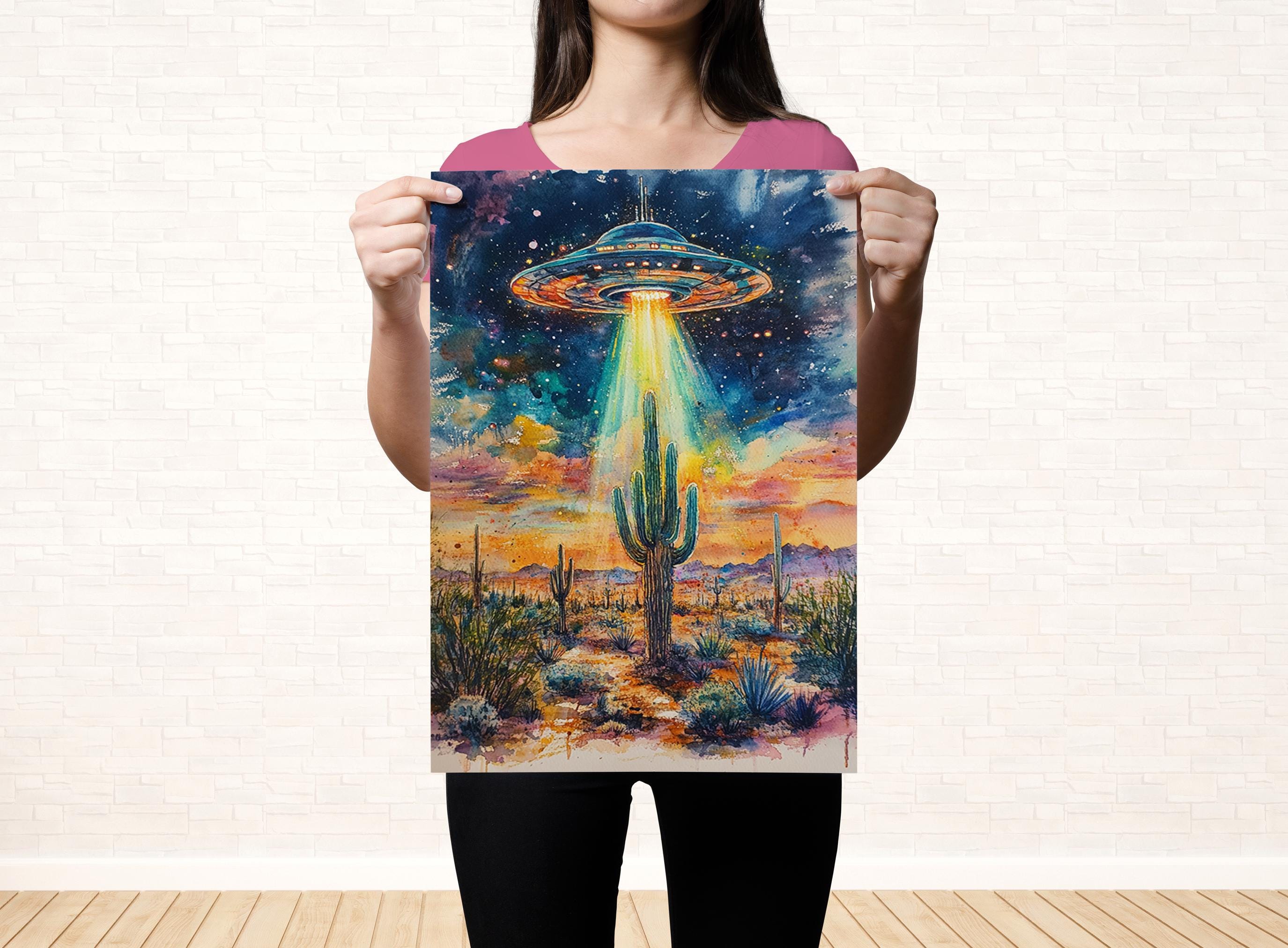 A woman holds up a poster featuring a vibrant painting of a desert landscape with a large alien spacecraft hovering above a cactus.