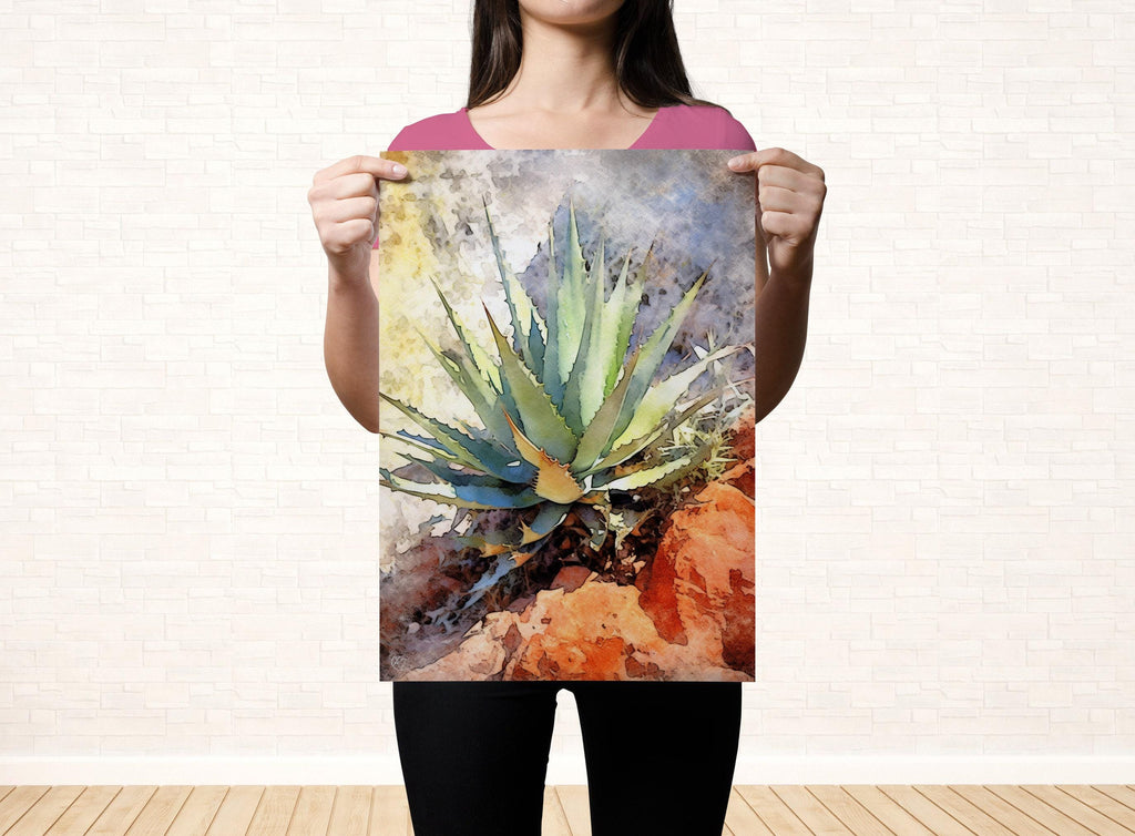 A woman holds up a framed print of a colorful, abstract painting of a cactus plant against a white brick wall.
