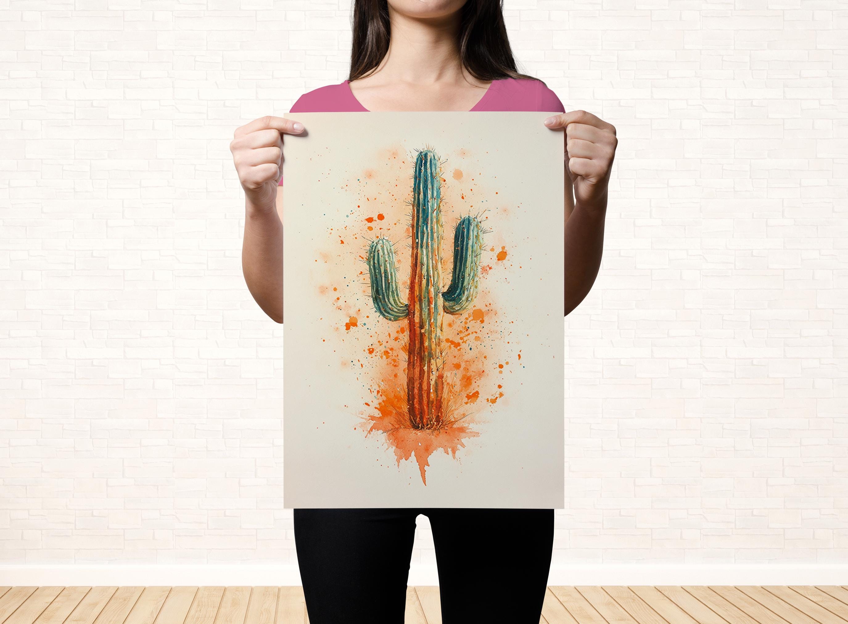 A woman is holding up a framed print of a cactus with a watercolor effect, featuring a vibrant mix of orange and green colors.