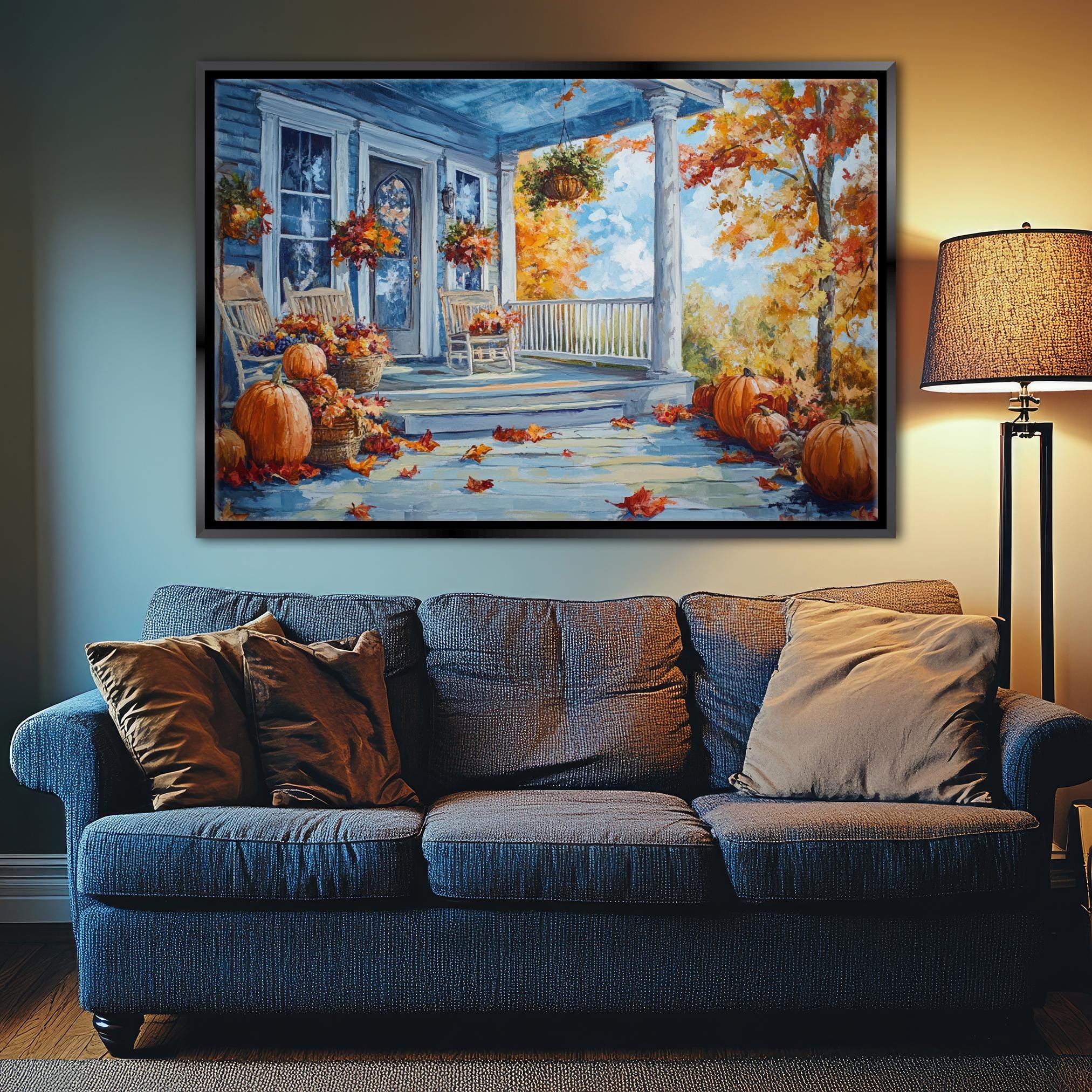 a large painting of a porch with a blue house, a white door, and a porch with a railing. The porch is decorated with pumpkins and autumn leaves, creating a cozy and festive atmosphere. The painting is hung on the wall above a blue couch, which is adorned with pillows. The overall scene conveys a sense of warmth and comfort, with the colors and details of the painting complementing the cozy setting of the living room.