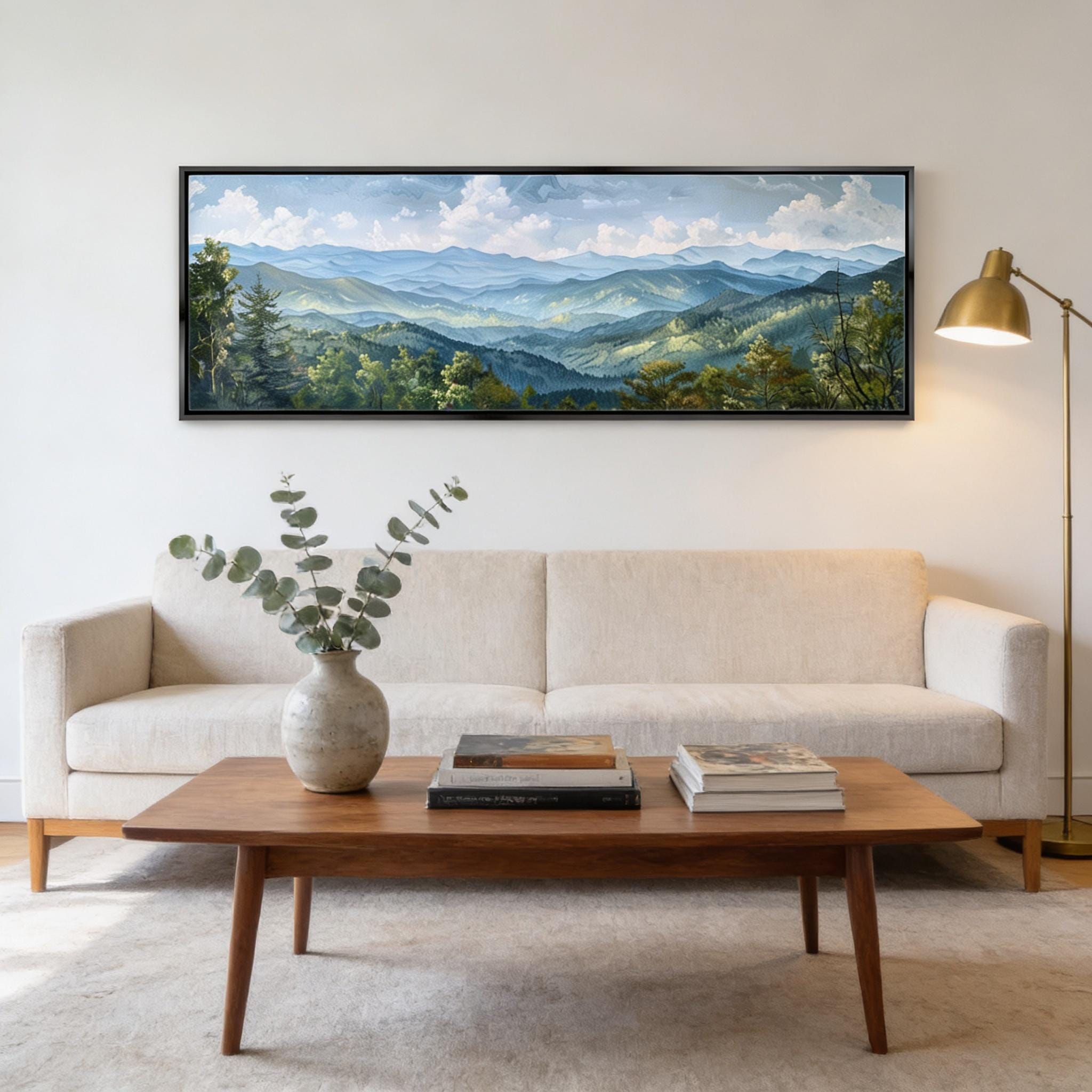 a living room with a large painting of a mountain landscape hanging on the wall above a wooden coffee table. The coffee table has a vase of greenery and a stack of books on it. The room has a white sofa and a lamp, creating a cozy and inviting atmosphere.
