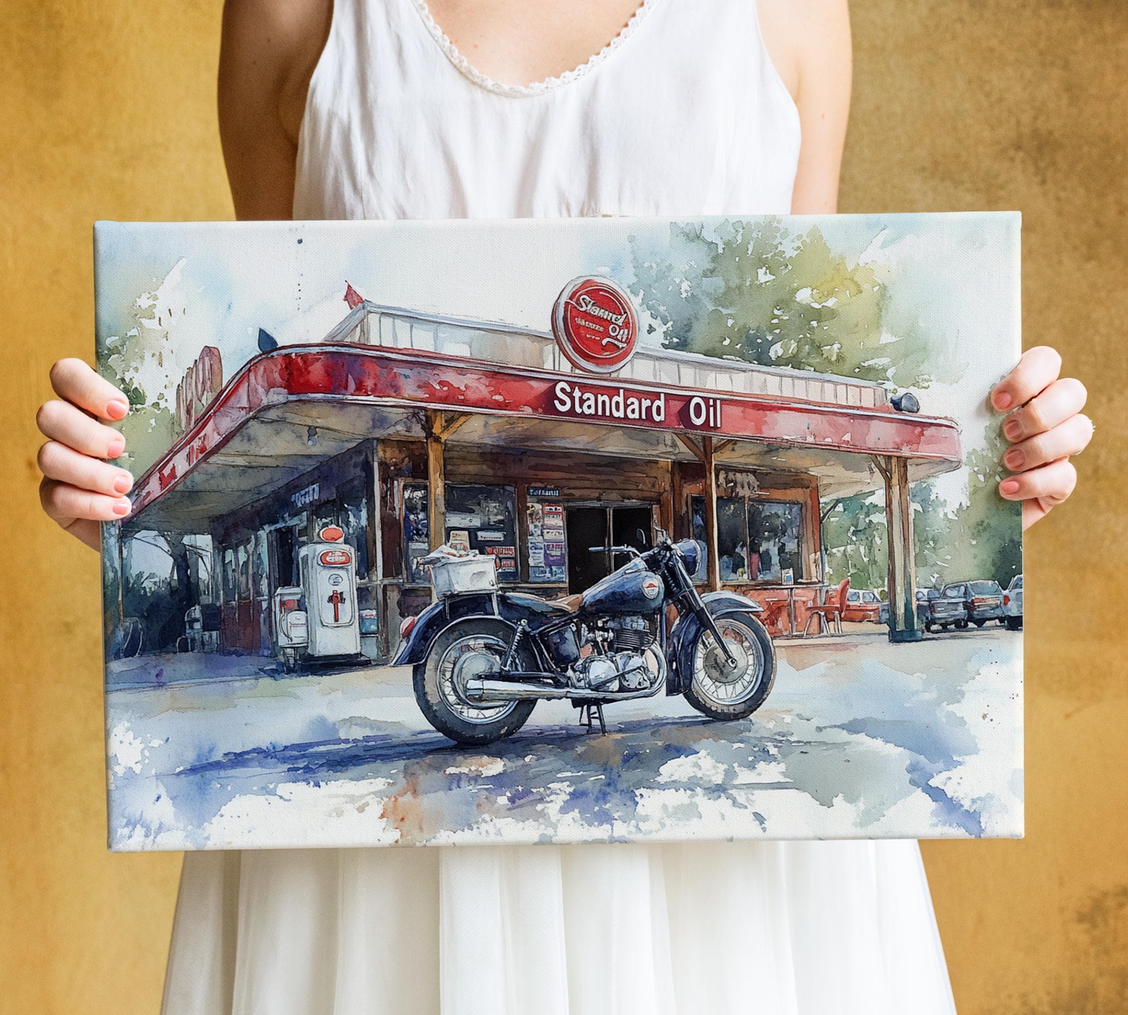 A person is holding a framed painting of a classic motorcycle parked in front of a vintage gas station.