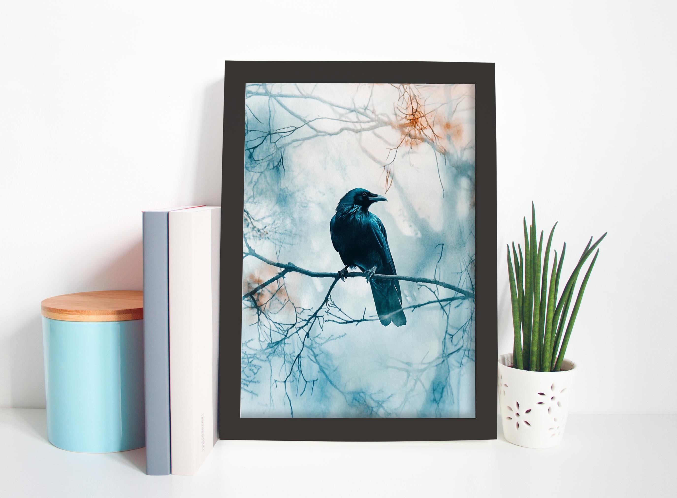 a framed photograph of a black crow perched on a branch, with a blue and white background. The photograph is displayed on a white shelf, accompanied by a blue book, a white vase with green plants, and a white ceramic container.