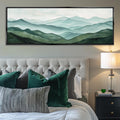 a serene bedroom scene with a large painting of a mountain landscape hanging above a tufted white headboard. The painting features a blend of green and gray tones, creating a calming and natural ambiance. The room is furnished with a tufted white headboard, a nightstand with a lamp, and a bed adorned with green and gray pillows.