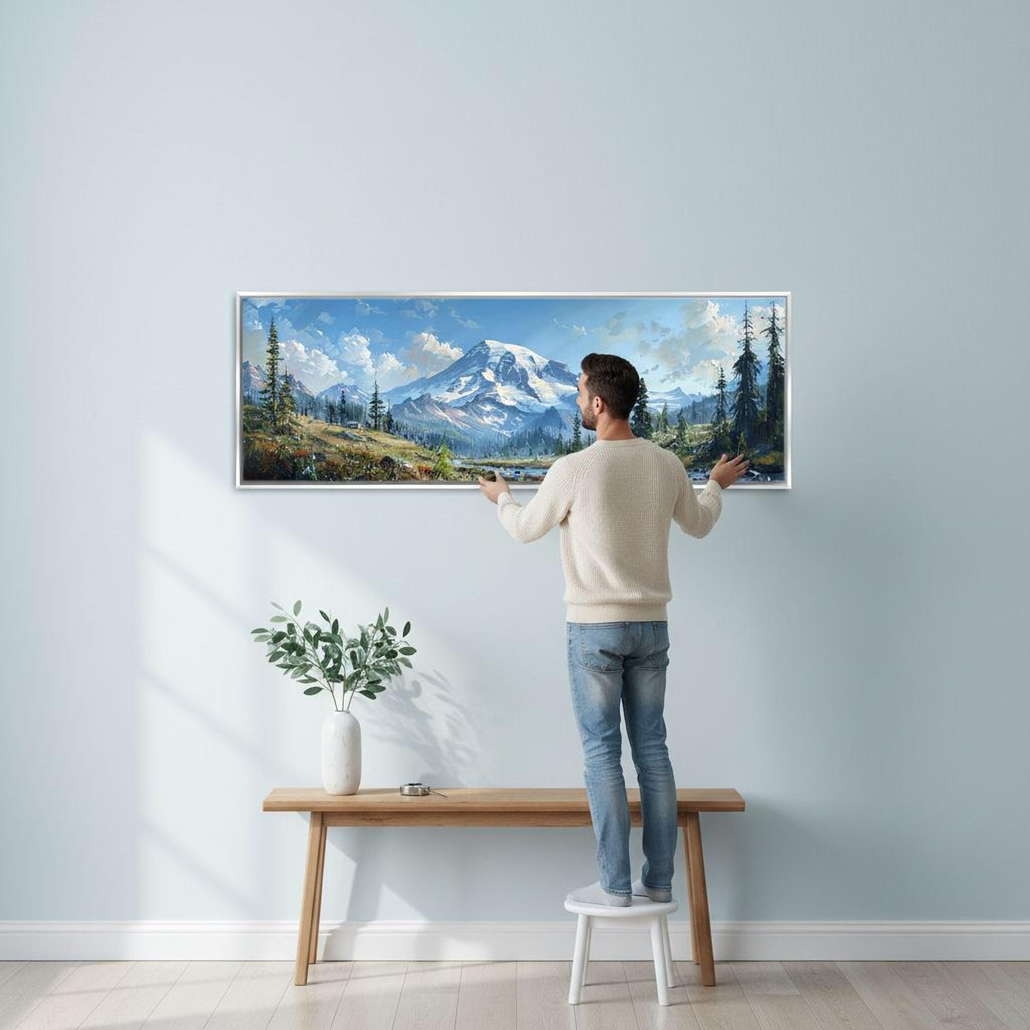 A man is standing on a stool and hanging a large landscape painting on a wall in a room with a wooden bench and a potted plant nearby.