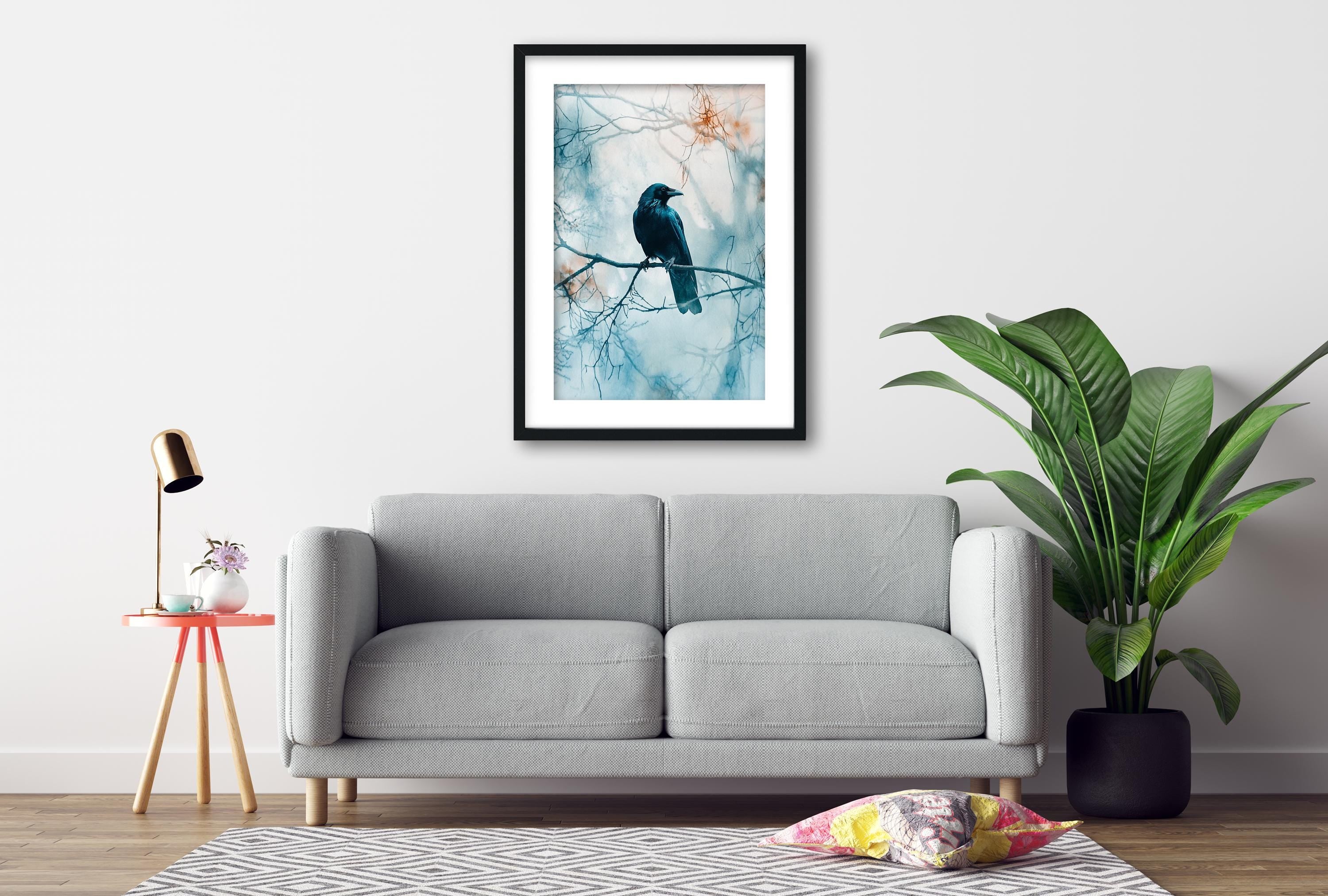 a gray sofa, a framed picture of a black bird perched on a branch, a small table with a lamp, and a potted plant.