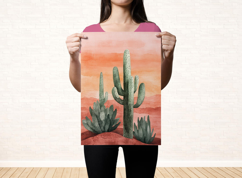 A woman is holding up a watercolor painting of a desert scene with cacti and a sunset in the background.