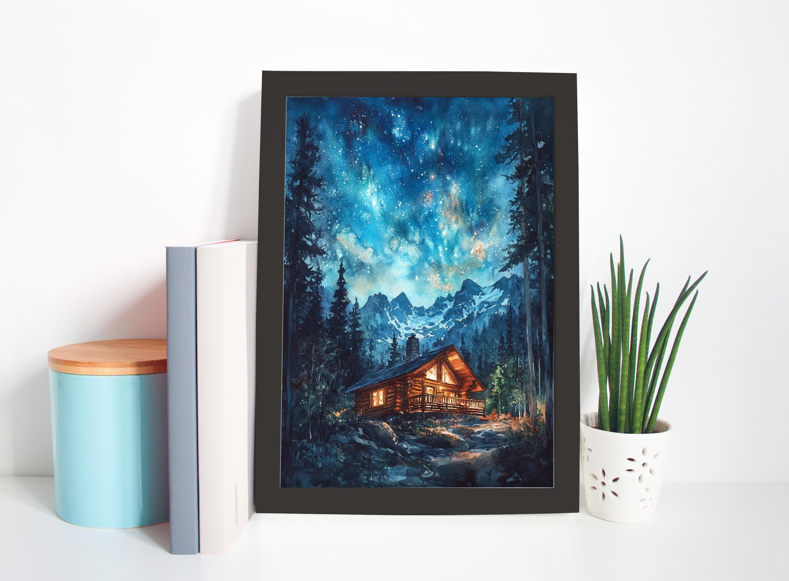 a framed painting of a cabin in a forest, with a starry night sky in the background. The painting is displayed on a white wall, accompanied by a blue container, a white vase with a plant, and a stack of books.