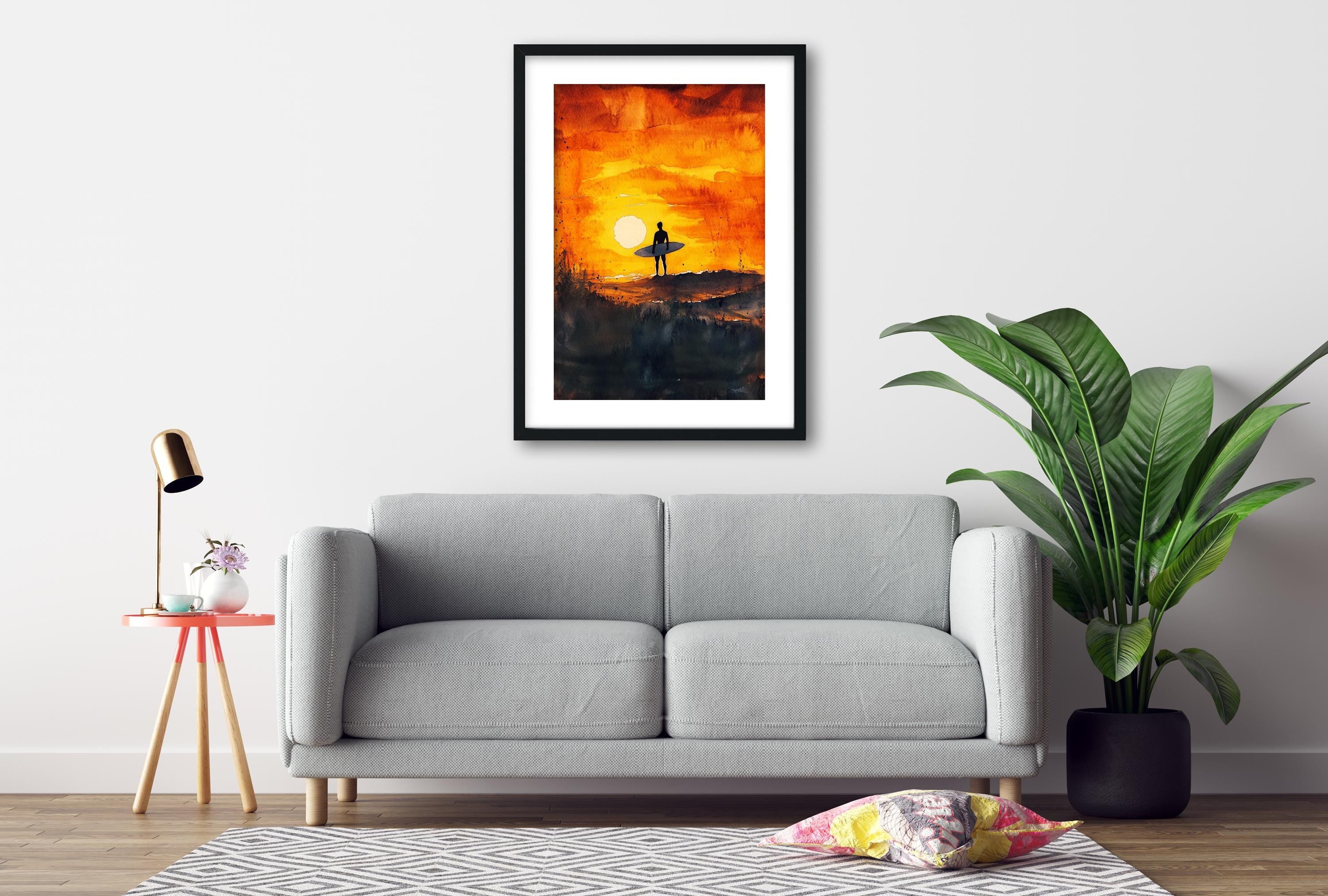a living room with a gray sofa, a framed painting of a surfer on a beach at sunset, a potted plant, and a small table with a lamp.